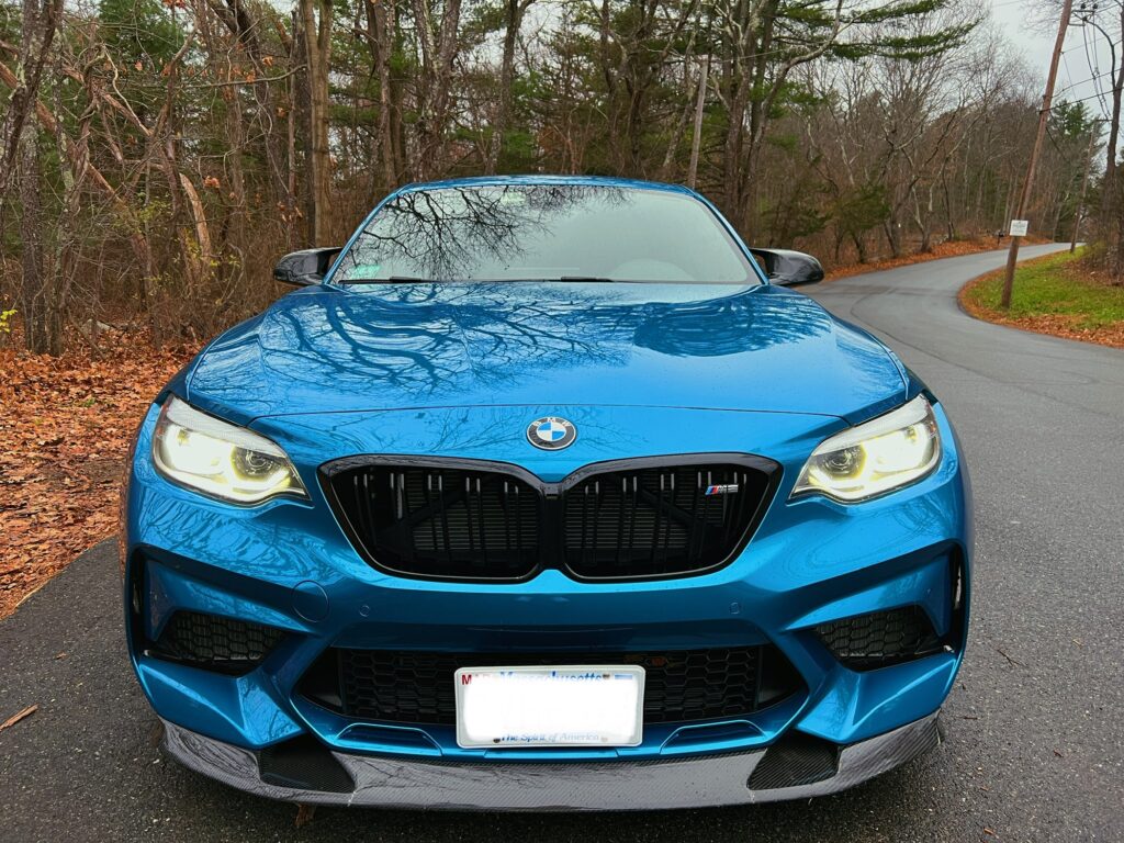 BMW M2 in vibrant blue color, showcasing aggressive front grille and sleek design, parked on a winding road surrounded by autumn foliage, highlighting Schneller BMW's focus on performance and quality service.