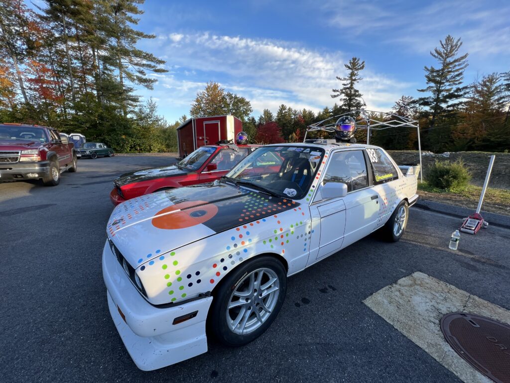 BMW race car with colorful dot patterns and a helmet on the roof, parked in a service area surrounded by trees and other vehicles.