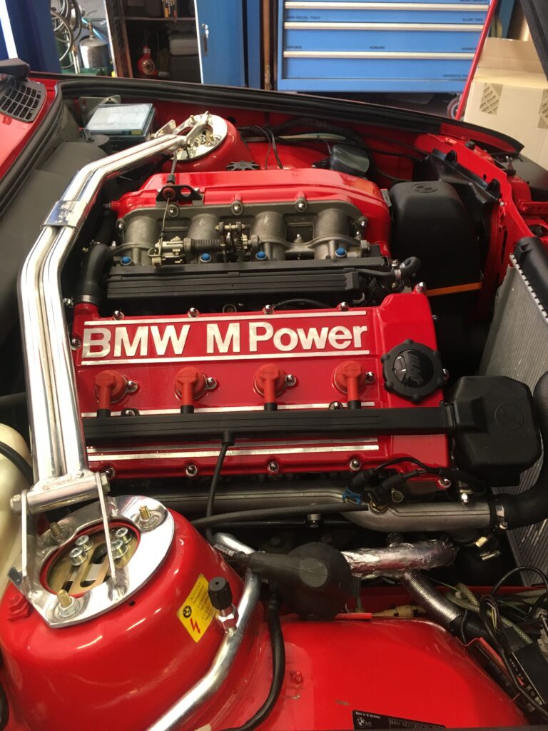 BMW M Power engine with red valve cover and polished intake manifold, showcased in a professional automotive workshop setting.