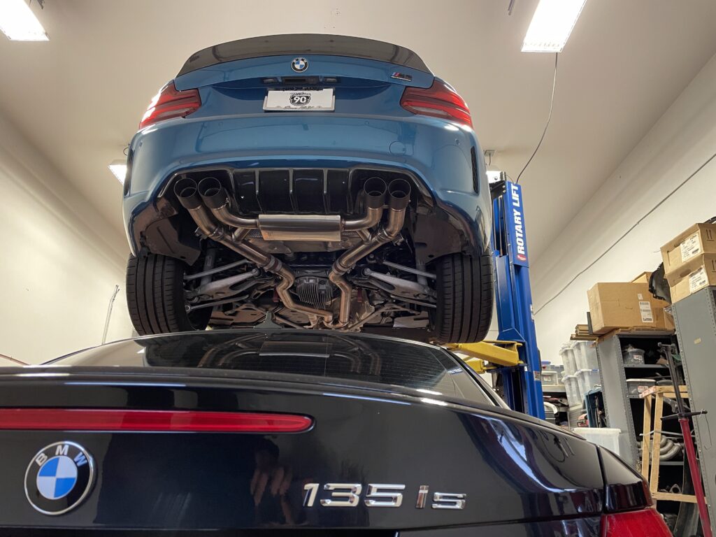 BMW vehicles in service at Schneller BMW, showcasing a blue BMW M2 on a lift above a black BMW 135is, emphasizing the facility's expertise in BMW maintenance and repairs.