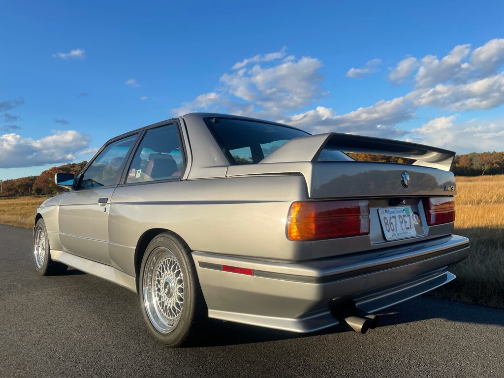 BMW E30 M3 coupe parked on a scenic road, showcasing its iconic design, chrome wire wheels, and rear spoiler, with a backdrop of autumn foliage and blue sky, reflecting the passion and expertise of Schneller BMW in servicing classic BMW vehicles.