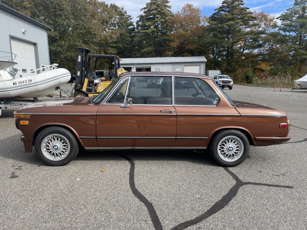 Classic BMW 2002 in brown parked outside Schneller BMW service facility, showcasing vintage design and attention to detail, surrounded by automotive equipment and trees in the background.