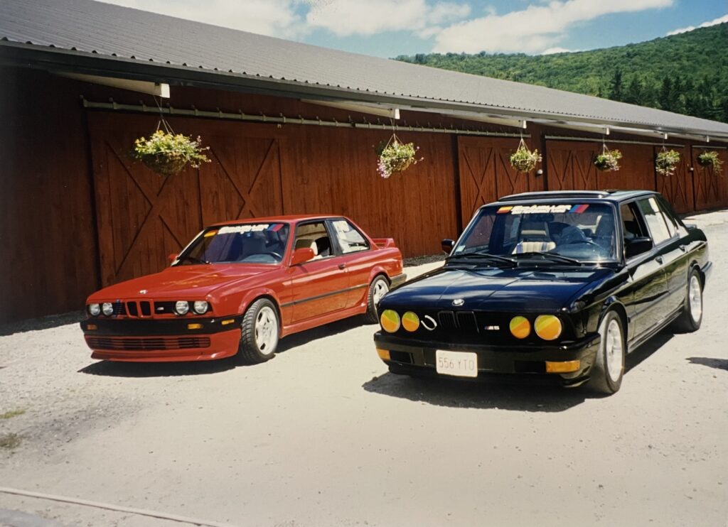 Red and black BMW cars parked in front of wooden barn, showcasing classic BMW models with distinctive features and aftermarket modifications, relevant to BMW diagnostic services.