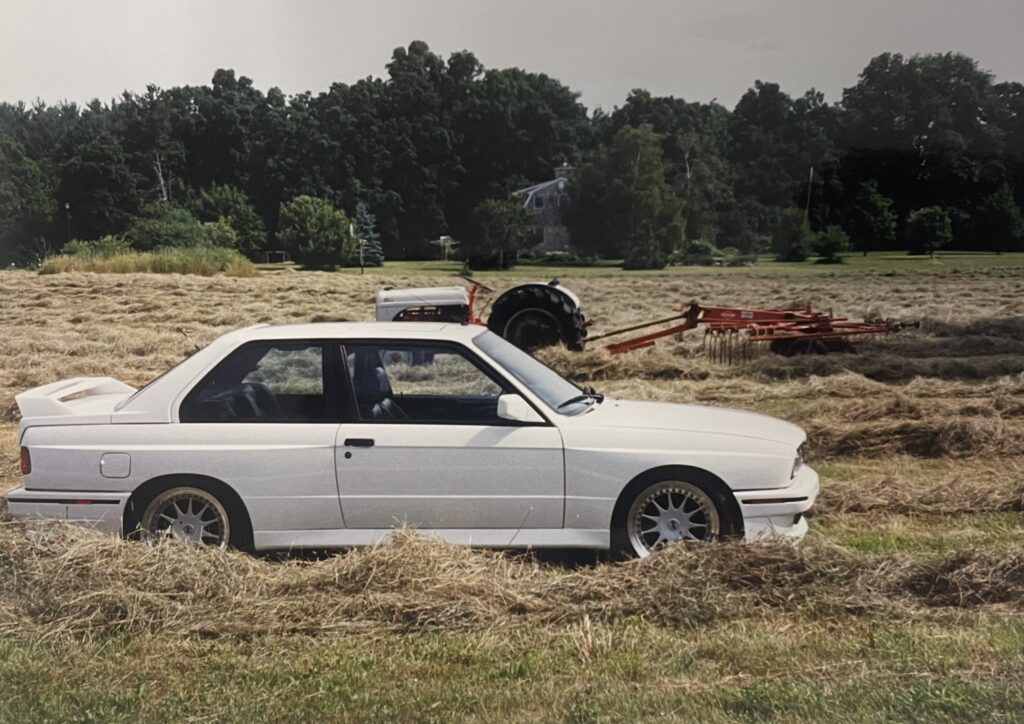 White BMW E30 coupe parked in a field of hay, with a tractor and trees in the background, reflecting the brand's connection to performance and automotive culture.