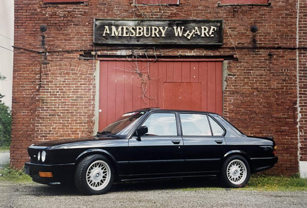 Black BMW parked in front of a rustic red brick building with an "Amesbury Wharf" sign, highlighting the vehicle's classic design and the facility's focus on BMW service and culture.