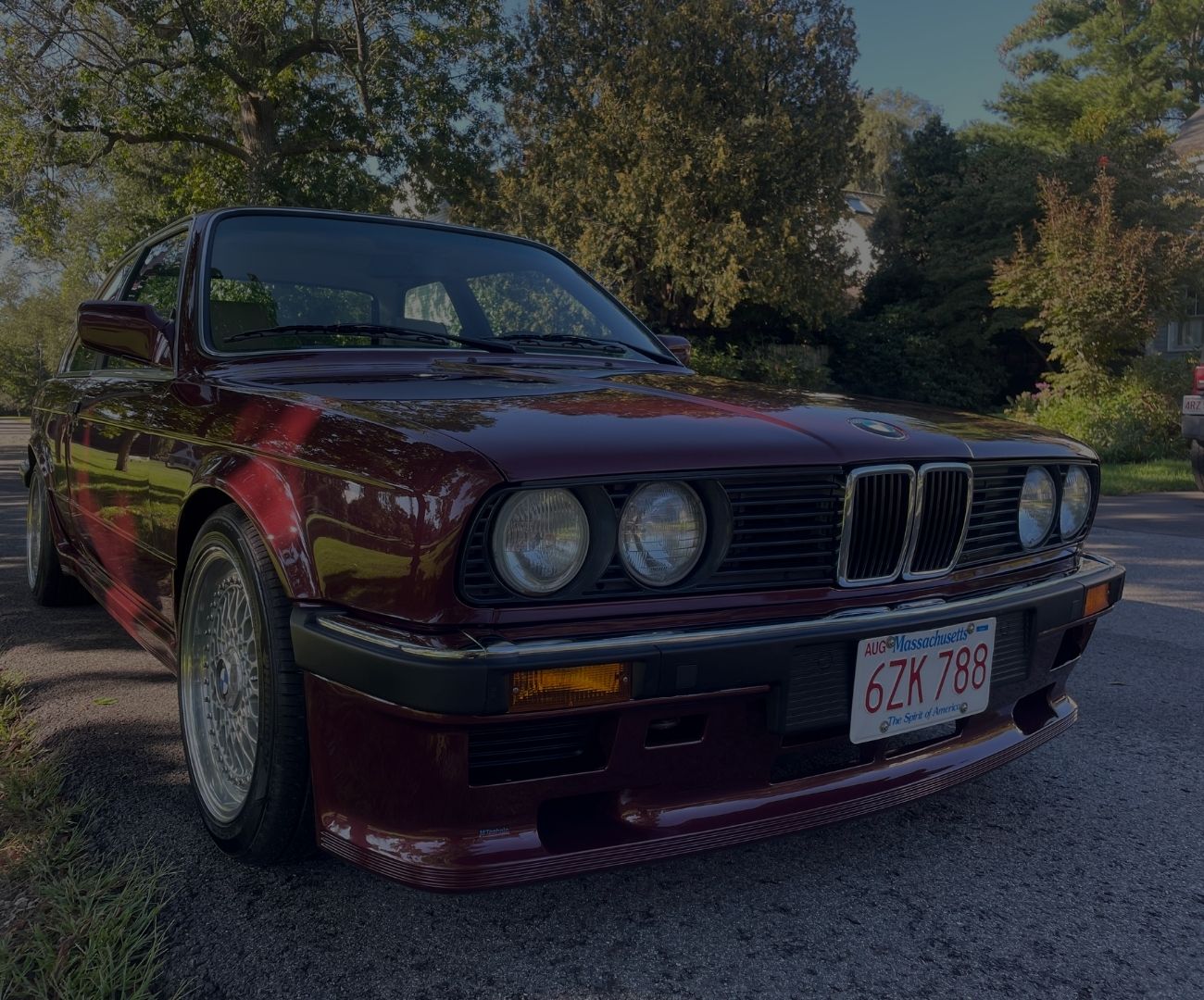 BMW E30 in burgundy color, showcasing classic design and detailed alloy wheels, parked on a tree-lined street, symbolizing Schneller BMW's expertise in E30 restorations and performance builds.